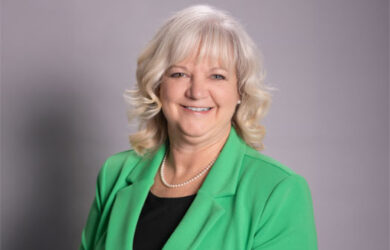A woman with shoulder-length blonde hair, wearing a green blazer, black top, and pearl necklace, smiles at the camera in front of a plain gray background.