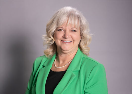 A woman with shoulder-length blonde hair, wearing a green blazer, black top, and pearl necklace, smiles at the camera in front of a plain gray background.