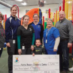 A group of adults and one child stand indoors in front of a colorful indoor playground, holding a large check for $2,000 made out to the Kenny Rogers Childrens Center. Everyone is smiling, and the date on the check is 12/13/19.