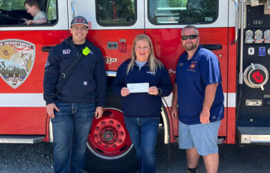 Three adults stand in front of a fire truck; one holds a check. The fire truck has “Lake City Fire Department” written on it. A child sits inside the truck.