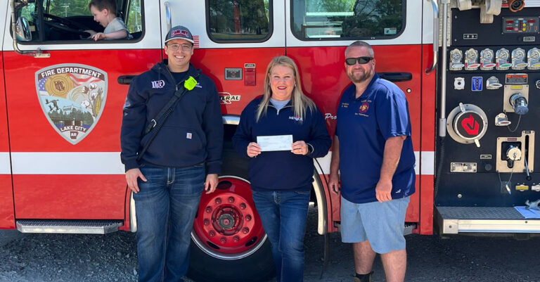 Three adults stand in front of a fire truck; one holds a check. The fire truck has “Lake City Fire Department” written on it. A child sits inside the truck.