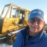 A man in sunglasses and a blue cap stands outdoors, smiling in front of a yellow Caterpillar bulldozer. The ground is covered in patches of snow, and hes wearing a blue jacket.