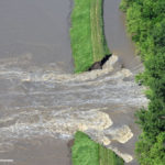 Aerial view of a river overflowing, breaching a grassy levee and flooding a nearby area. The brown floodwaters rush towards a green forest on the right side of the image.