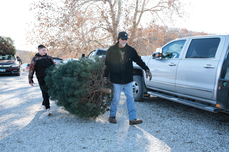 Two people carrying a Christmas tree near parked cars. One person wears a jacket and sunglasses, the other a patterned sweater. The background has leafless trees and a clear sky.