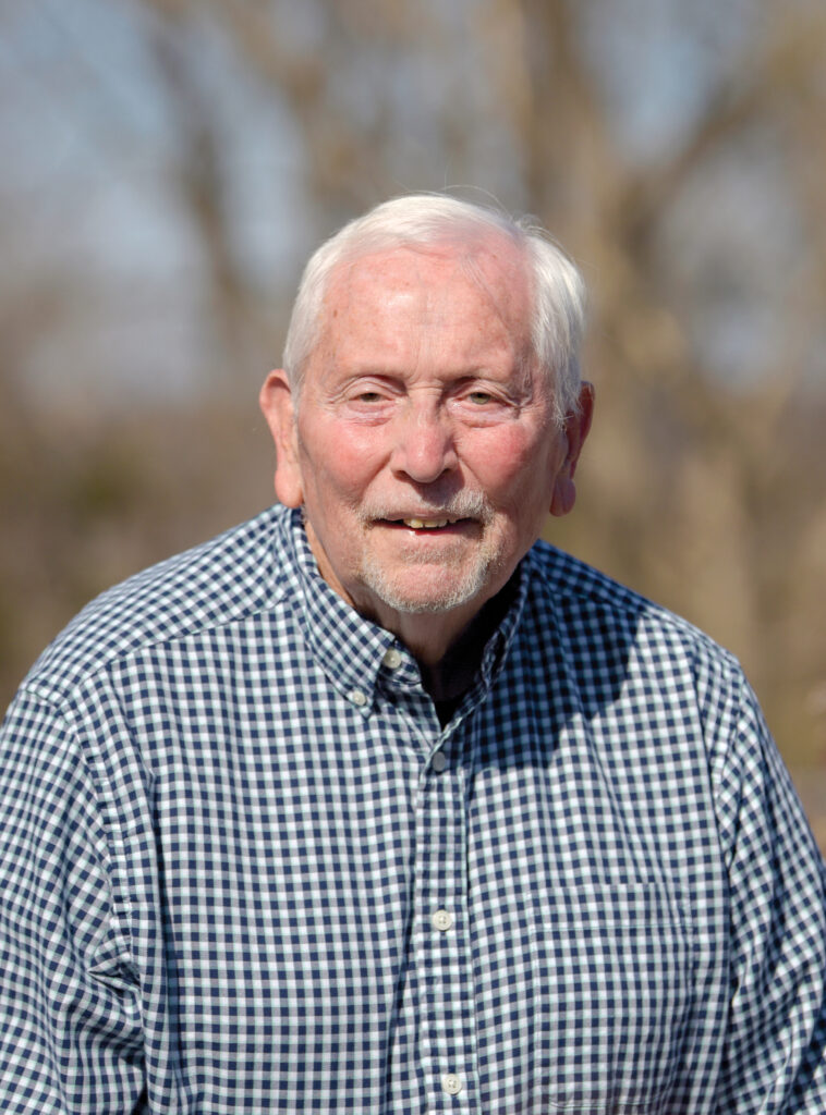 Lowell Mohler with short white hair and a white beard smiles at the camera while wearing a blue and white checkered shirt. The background is outdoors and blurred with trees.