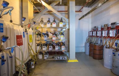 A lubricant storage room filled with various labeled chemical containers, hoses, and barrels. The walls are painted white, and the floor is concrete. Shelving units hold multiple small containers with different colored labels.