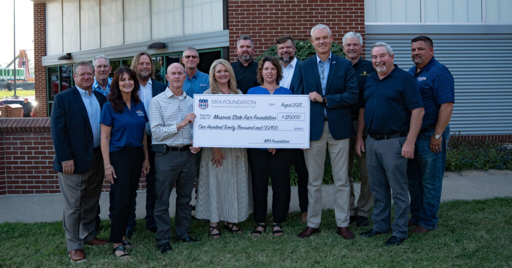 A group of people stands outside a brick building, smiling and holding an oversized check from the MFA Foundation for $120,000, made out to the Arkansas State Fair Foundation.