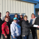 A group of nine people stand outside a building. Two individuals at the front are shaking hands and holding a check. Everyone is smiling, and theyre dressed in casual, warm clothing. The building has a metal facade and a blue roof.
