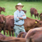 A farmer wearing a wide-brimmed hat and plaid shirt stands in a lush green field, surrounded by a herd of brown cows. He is smiling with arms crossed, exuding a sense of contentment and pride in his pastoral setting.