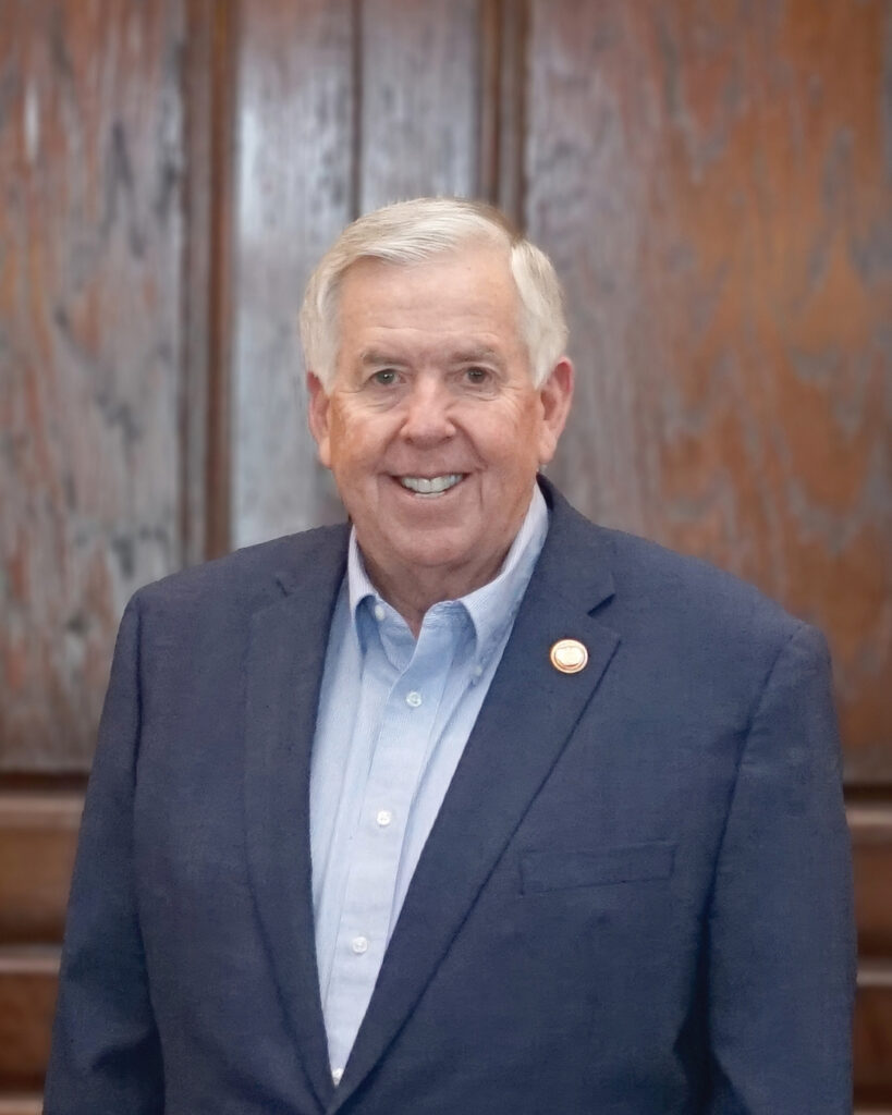 Former Missouri Governor Mike Parson wearing a blue suit jacket, light blue dress shirt, and a pin on his lapel stands in front of a wooden paneled background.