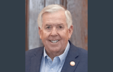 Former Missouri Governor Mike Parson wearing a blue suit jacket, light blue dress shirt, and a pin on his lapel stands in front of a wooden paneled background.