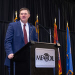 A man in a blue suit stands at a podium with an MFA Oil logo, speaking into a microphone. Behind him are several flags against a black curtain backdrop.