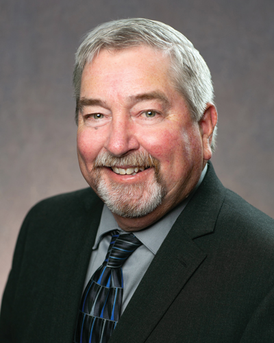 Portrait of Mike Moreland District 5 West Central Bord Member man with gray hair and beard wearing a dark suit, blue striped tie, and gray shirt against a gray background.