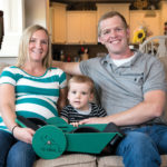 A smiling couple sits on a couch with their young child. The woman wears a striped shirt and holds a green toy labeled The Frog. The man in a gray shirt sits to their right. The living room background features a kitchen and sunflowers.