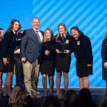 A group of people on stage wearing blue jackets and smiling. One person holds a trophy. A blue backdrop with diagonal stripes is behind them.