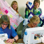 Four women are engaged in quilting activities. Two women in the foreground are sewing on machines, while two women in the background hold up colorful quilts, smiling. The scene appears lively and cheerful.