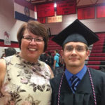 A smiling graduate in cap and gown stands beside a woman in a floral top. They are inside a gymnasium with red and black bleachers and championship banners on the walls.