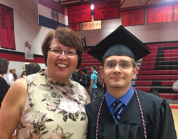 A smiling graduate in cap and gown stands beside a woman in a floral top. They are inside a gymnasium with red and black bleachers and championship banners on the walls.
