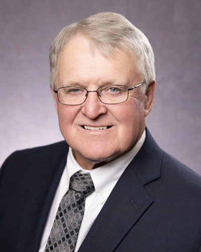 Portrait of Doyle Oehl District 4 Southeast board member man with short gray hair and glasses, wearing a dark suit, white shirt, and patterned tie, smiles at the camera against a plain background.