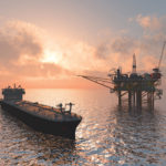 A large oil tanker is positioned near an offshore oil rig under a vivid sunset sky. The ocean reflects the colorful hues of the setting sun, creating a serene yet industrial scene.