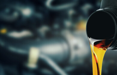Golden engine oil is being poured from a black plastic bottle, with a blurred background showing parts of a car engine.