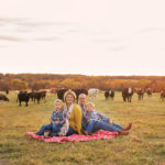 A family of four sits on a red patterned blanket in a grassy field. Two children, a man, and a woman are smiling at the camera. Cows are grazing in the background under a cloudy sky with a warm sunset glow.