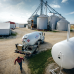 A truck with a propane tank is parked at a farm with large grain silos. A person walks nearby on a gravel path. In the background are blue skies, a field, and several farm buildings.