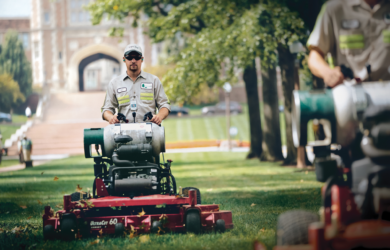 A person wearing a cap and sunglasses operates a lawnmower on a grassy area surrounded by trees. Another individual is partially visible in the background, also using a lawnmower. A large building with arches is in the distance.