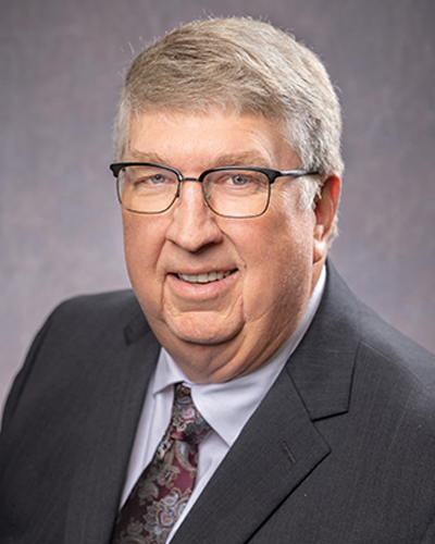 A man wearing glasses and a gray suit with a patterned tie smiles at the camera against a neutral background.