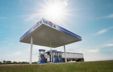 A solitary gas station with MFA Oil branding stands under a bright sun in a vast grassy field. A long truck is parked beside the pumps. The sky is clear and blue, casting long shadows on the surrounding landscape.