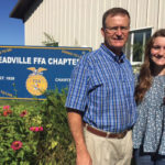 A man and a woman stand in front of a sign for Meadville FFA Chapter. The man wears a blue plaid shirt and glasses, while the woman has long brown hair and is wearing a blue patterned blouse. Bright flowers are in the foreground.