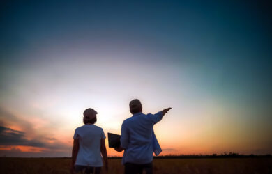 Two people in hats stand in a field at sunset; one holds a laptop while the other points toward the horizon, silhouetted against a colorful sky.