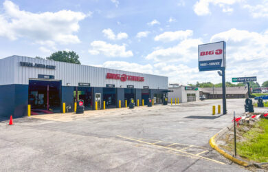 A Big O Tires auto shop with multiple service bays and a large sign in front. The lot is mostly empty under a partly cloudy sky, and an Enterprise building is visible in the background.