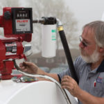 A person with gray hair and a beard is operating a red fuel transfer pump, attaching a hose to the device. The pump has visible dials and a display. A filter labeled Bio* is connected to the setup. The background is slightly foggy.