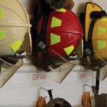 Firefighter helmets hanging on hooks, labeled for roles such as ASST. CHIEF and CAPTAIN. Helmets are in various colors, including white, red, and yellow, with full firefighting gear underneath them.