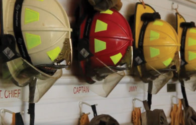 Firefighter helmets hanging on hooks, labeled for roles such as ASST. CHIEF and CAPTAIN. Helmets are in various colors, including white, red, and yellow, with full firefighting gear underneath them.