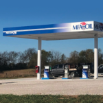 A rural gas station with MFA Oil branding, featuring two pumps under a blue and white canopy. A pickup truck towing a large white tank is parked nearby, with trees and a clear blue sky in the background.