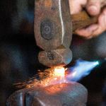 A close-up of a blacksmiths hand holding a hammer striking a hot, glowing piece of metal on an anvil, with sparks flying. The image captures the intense heat and energy of the metalworking process.