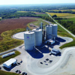 Aerial view of a large agricultural silo complex in a rural landscape. Surrounding fields with greenery and brown plots. Roads and a small building are nearby, with several parked vehicles. The horizon is visible in the background.