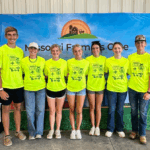 Seven young adults stand side by side, smiling, and wearing matching bright yellow T-shirts in front of a banner that reads Missouri Farmers Care with a farm graphic at the top.