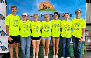 Seven young adults stand side by side, smiling, and wearing matching bright yellow T-shirts in front of a banner that reads Missouri Farmers Care with a farm graphic at the top.