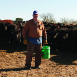 A person in a brown jacket and blue cap stands in front of a herd of cattle on a farm. They are holding a green bucket and wearing gloves, with a clear sky in the background.