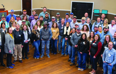 A large group of people, diverse in age and gender, standing together indoors. They are dressed in casual and business casual attire, many wearing name tags. The group is smiling, and the setting appears to be a conference or seminar room.