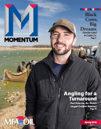A man wearing a cap and jacket smiles at the camera. Behind him, people work with nets in a large fish pond. The cover text includes Momentum, Angling for a Turnaround, and info about MFA Oil.