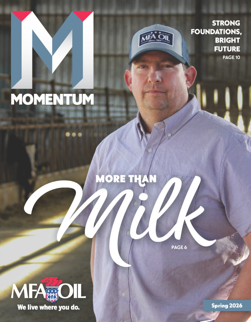A man wearing a blue MFA Oil cap and plaid shirt stands in a barn. The magazine cover reads Momentum: More Than Milk with the MFA Oil logo and Spring 2026 in the corner.