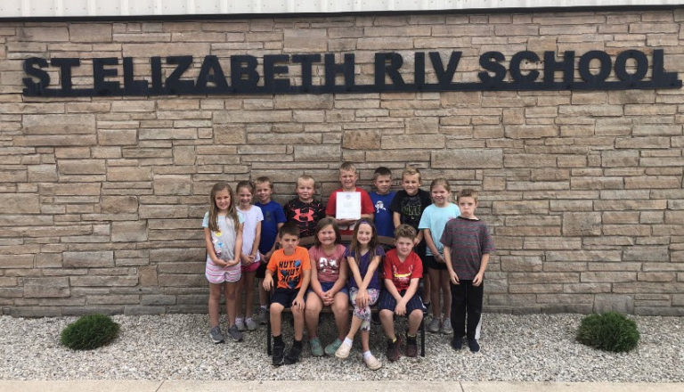 A group of children pose in two rows in front of a brick wall with ST. ELIZABETH R-IV SCHOOL on it. One child in the front row holds up a document. The kids are smiling and casually dressed.