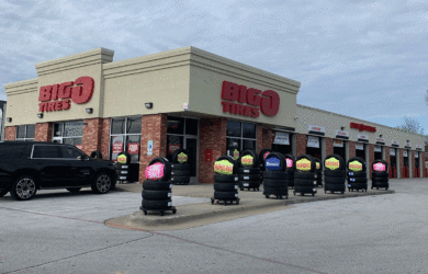 A Big O Tires store with a large parking lot in front. Several tire stacks with colorful sale signs are lined up outside. A black SUV is parked nearby. The sky is cloudy.