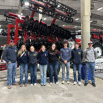 A group of nine people, wearing casual jeans and jackets, stand in front of a large agricultural machine at an indoor event. They pose for the photo with smiles. The setting appears industrial with exposed beams and concrete flooring.