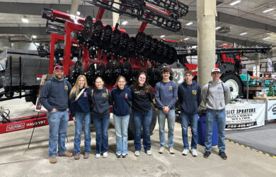 A group of nine people, wearing casual jeans and jackets, stand in front of a large agricultural machine at an indoor event. They pose for the photo with smiles. The setting appears industrial with exposed beams and concrete flooring.
