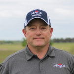A man wearing a dark gray MFA Oil Propane polo shirt and matching baseball cap stands outdoors in a field with a cloudy sky in the background.